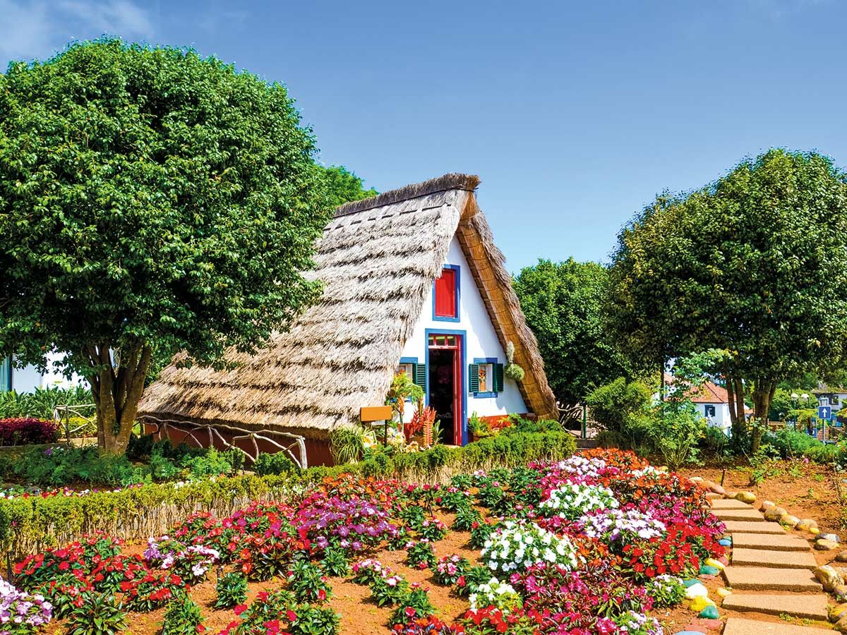 Traditionelles Santana Haus mit Strohdach, umgeben von bunten Blumenbeeten und grünen Bäumen unter blauem Himmel auf Madeira.