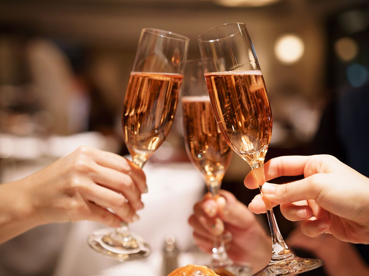 Nahaufnahme von drei Händen, die mit Gläsern voll roséfarbenem Sekt anstoßen, im Hintergrund unscharfe Lichter.