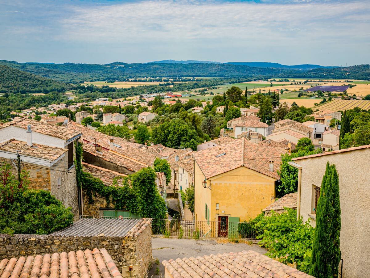 Malerisches Dorf in der Provence mit roten Dächern, grüner Vegetation und lila Lavendelfeldern im Tal unter blauem Himmel.