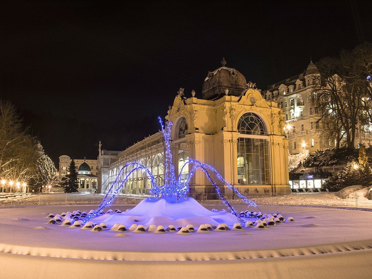 Nachtaufnahme eines historischen, beleuchteten Gebäudes mit schneebedecktem Brunnen und blauen LED-Lichtern in winterlicher Landschaft.