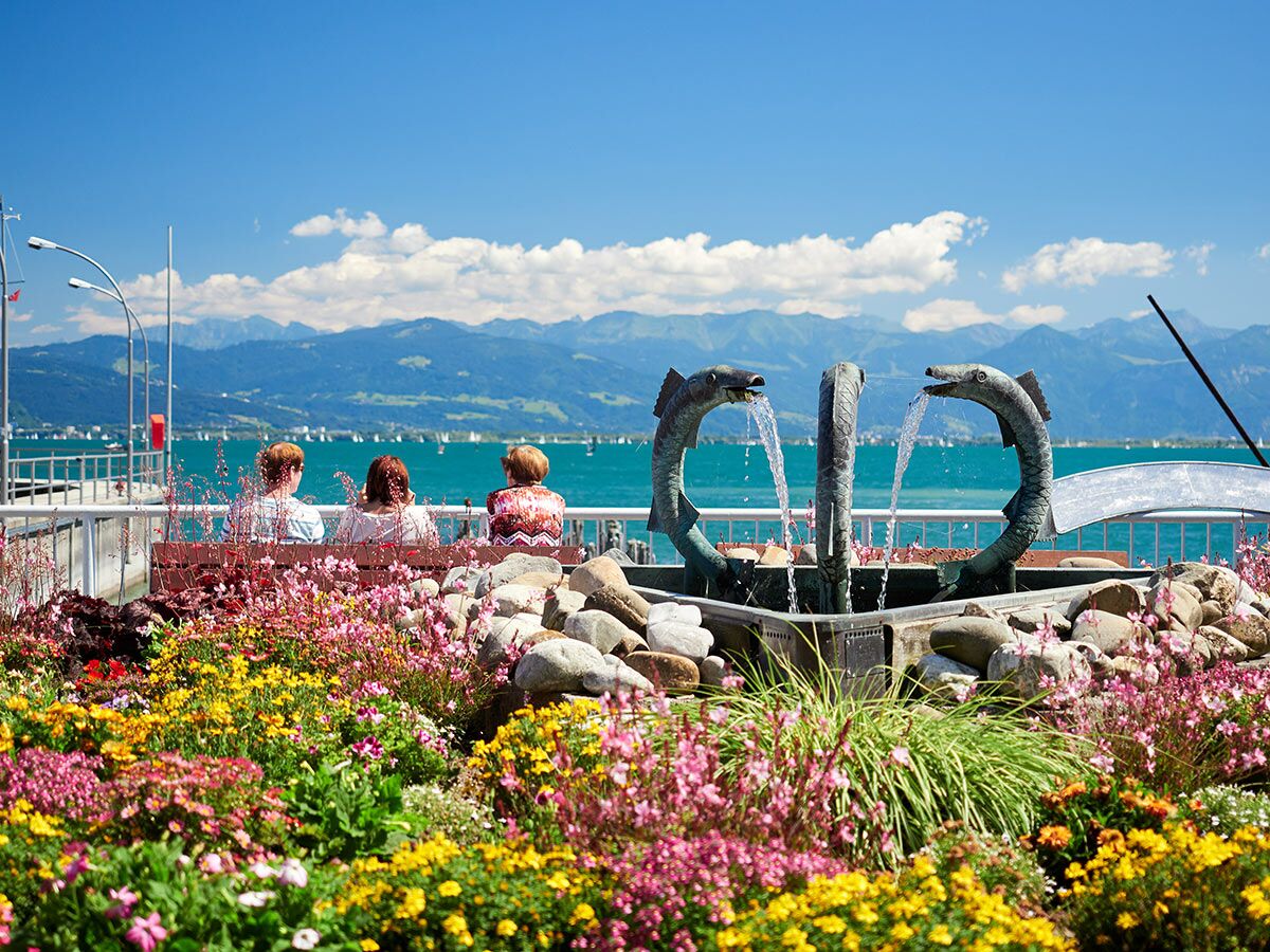 Drei Personen entspannen am Ufer des Sees mit Blumen, Brunnen und Bergpanorama im Sonnenschein.