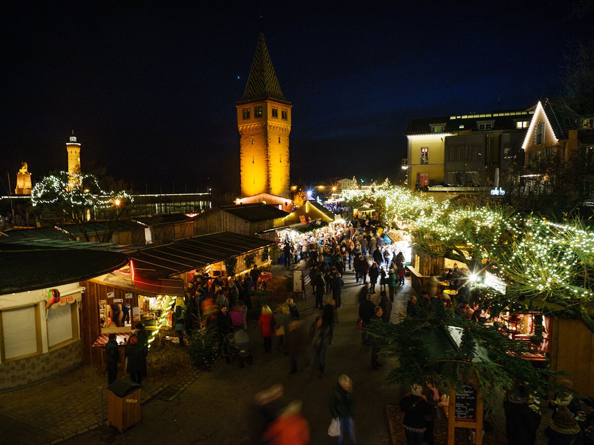 Festlicher Weihnachtsmarkt Lindau am Bodensee bei Nacht, mit beleuchteten Ständen, Menschen, Mangturm und Leuchtturm.