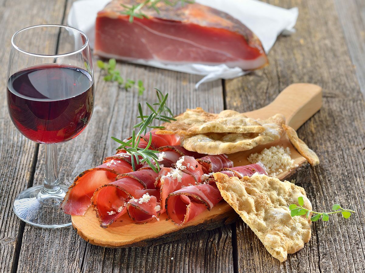 Glas Rotwein, Schinkenröllchen mit Käse und Kräutern, dazu Fladenbrot auf rustikalem Holzbrett serviert.