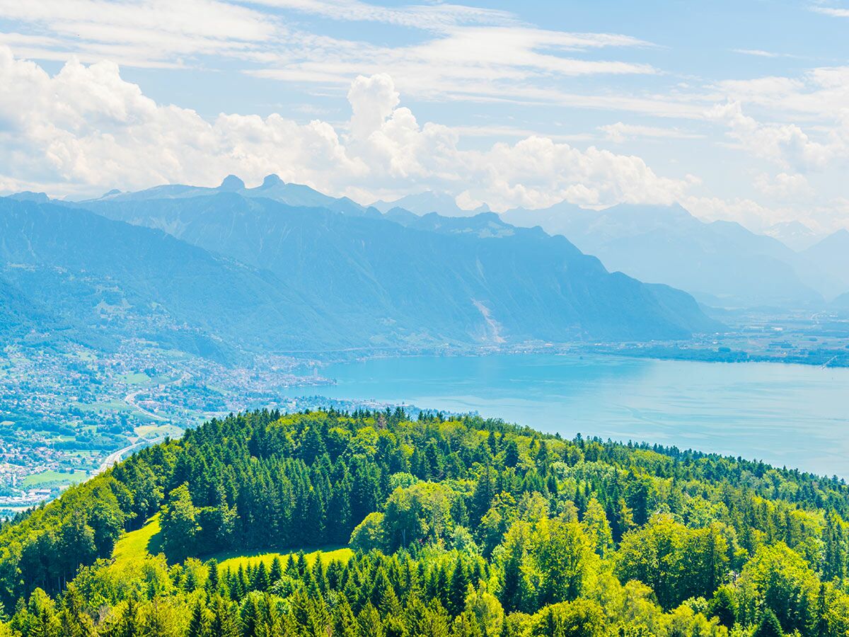 Panoramablick auf einen blauen Bergsee, umgeben von grünen Wäldern und majestätischen Alpenbergen unter blauem Himmel mit Wolken.