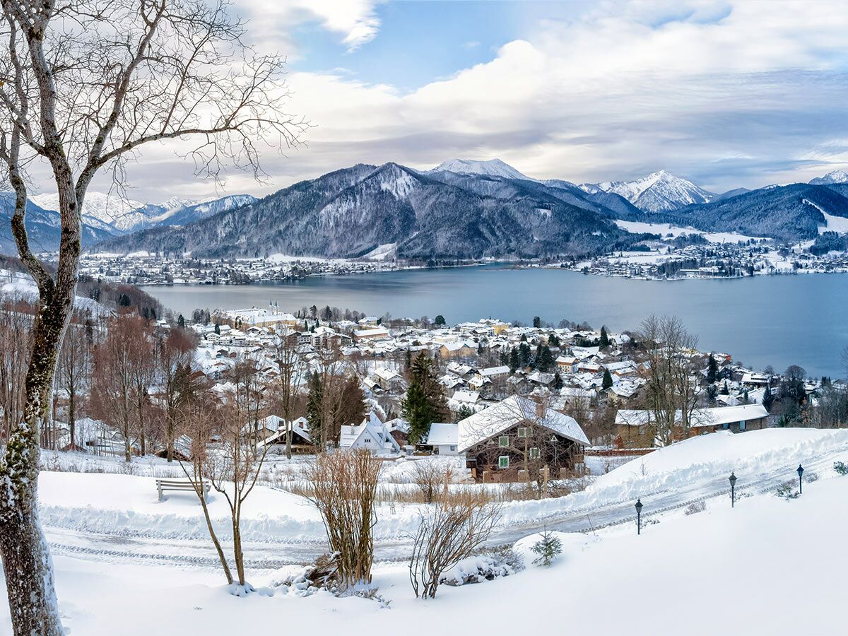 Panoramablick auf ein schneebedecktes Dorf am See, umgeben von verschneiten Alpen unter blauem Himmel.