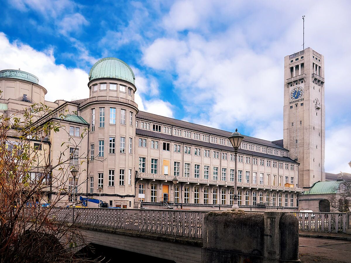 Ansicht des Deutschen Museums in München mit markantem grünem Kuppelbau und hohem Wettersäule-Turm unter blauem Himmel.