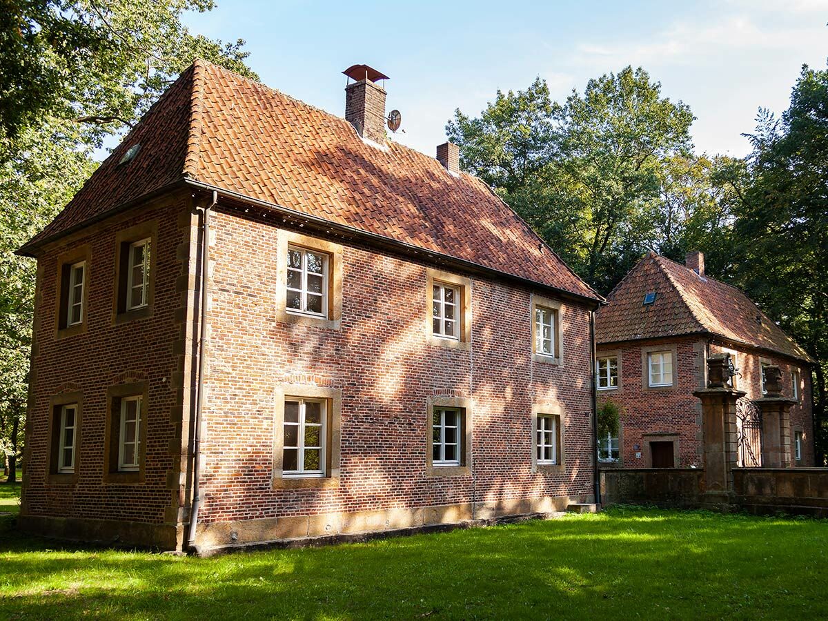 Zwei historische Backsteinhäuser mit roten Ziegeldächern und weißen Fenstern auf grünem Rasen, umgeben von Bäumen bei Sonnenschein.