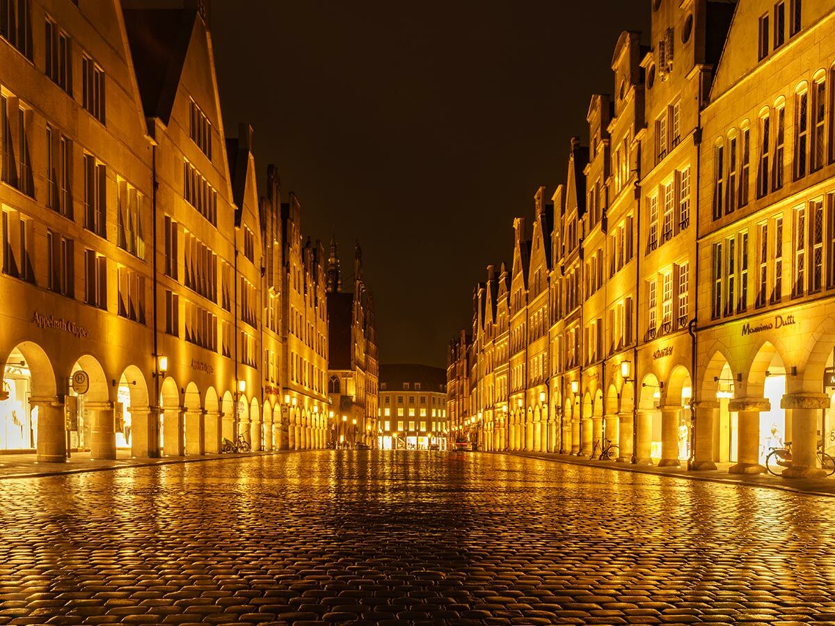 Der beleuchtete Prinzipalmarkt in Münster bei Nacht mit nasser Kopfsteinpflasterstraße, Arkaden und warmen Lichtreflexionen.