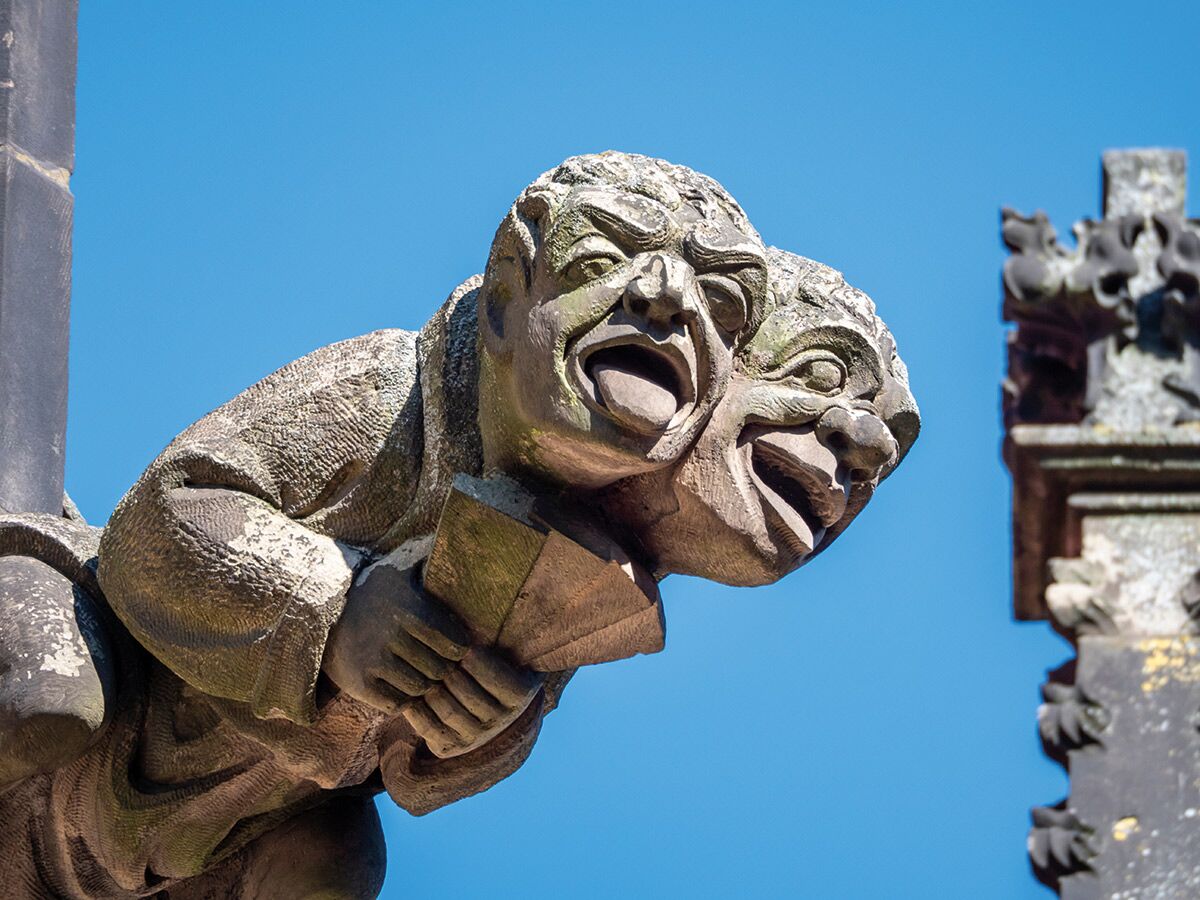 Detaillierte Aufnahme eines grotesken steinernen Gargoyles mit zwei schreienden Gesichtern und einem Buch vor blauem Himmel.