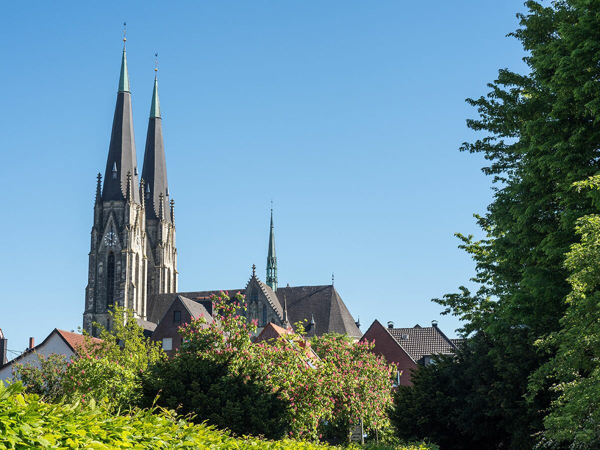 Gotische Kirche mit zwei hohen Türmen und einem kleineren Turm über Dächern und grünen Bäumen unter blauem Himmel.