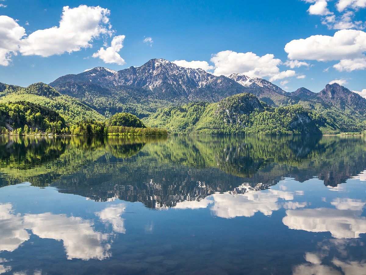 Ruhiger Bergsee mit majestätischen bewaldeten Bergen und schneebedeckten Gipfeln spiegelt blauen Himmel und weiße Wolken wider.