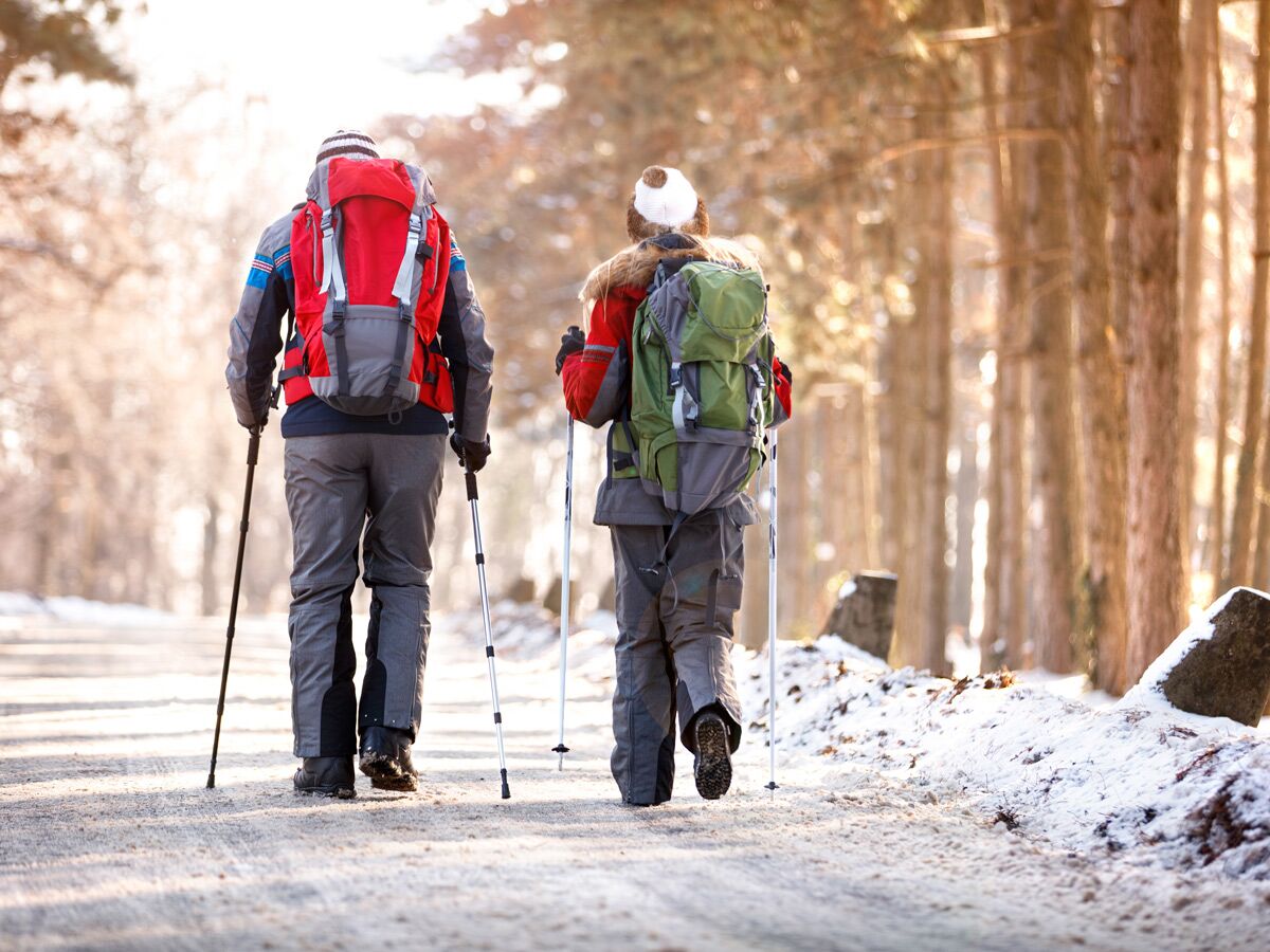 Zwei Wanderer mit Rucksäcken und Stöcken gehen auf einem schneebedeckten Waldweg in einer Winterlandschaft.