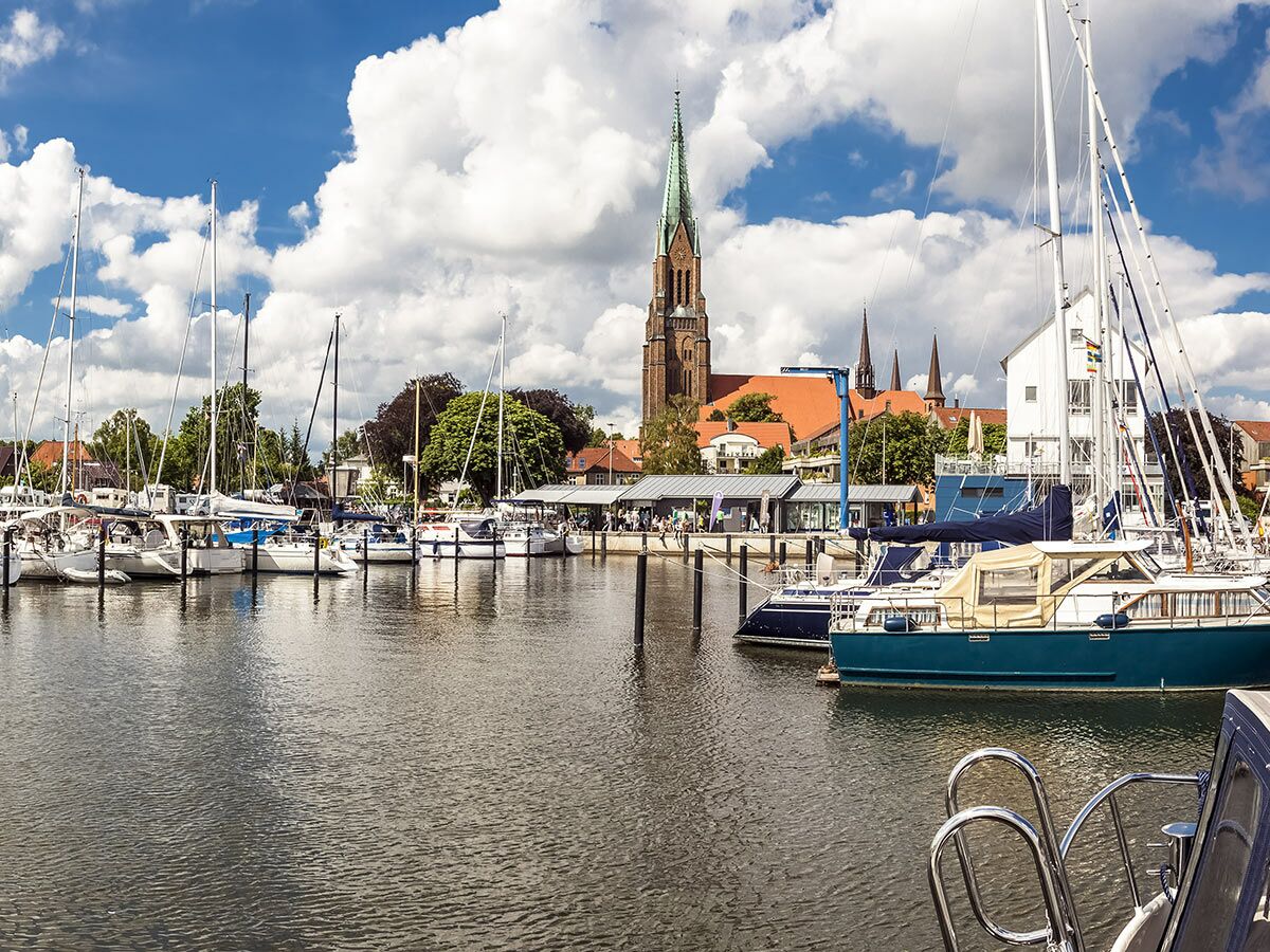 Malerischer Hafen mit vielen Segelbooten und Yachten, vor einer historischen Stadtkulisse mit hohen Kirchtürmen unter blauem Himmel.