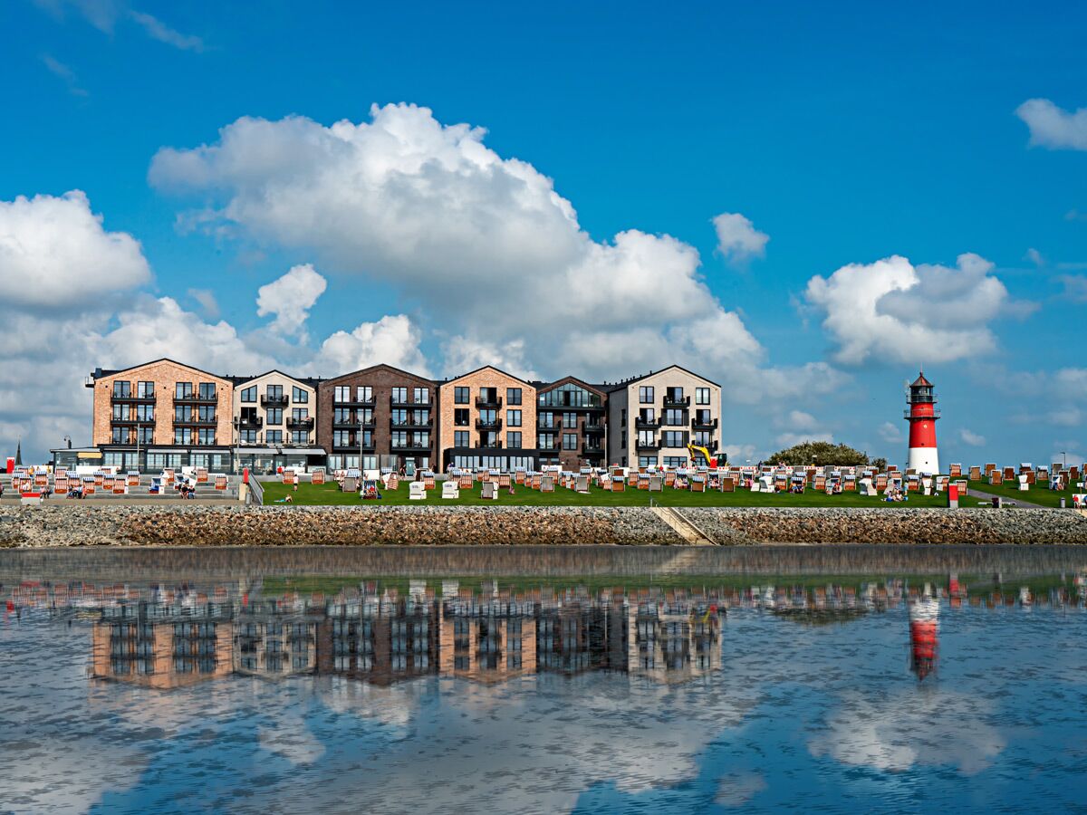 Modernes Küstenhotel mit Ziegelsteinfassaden und Balkonen, roter Leuchtturm, Strandkörbe an Promenade und Wasserreflexionen unter blauem Himmel.