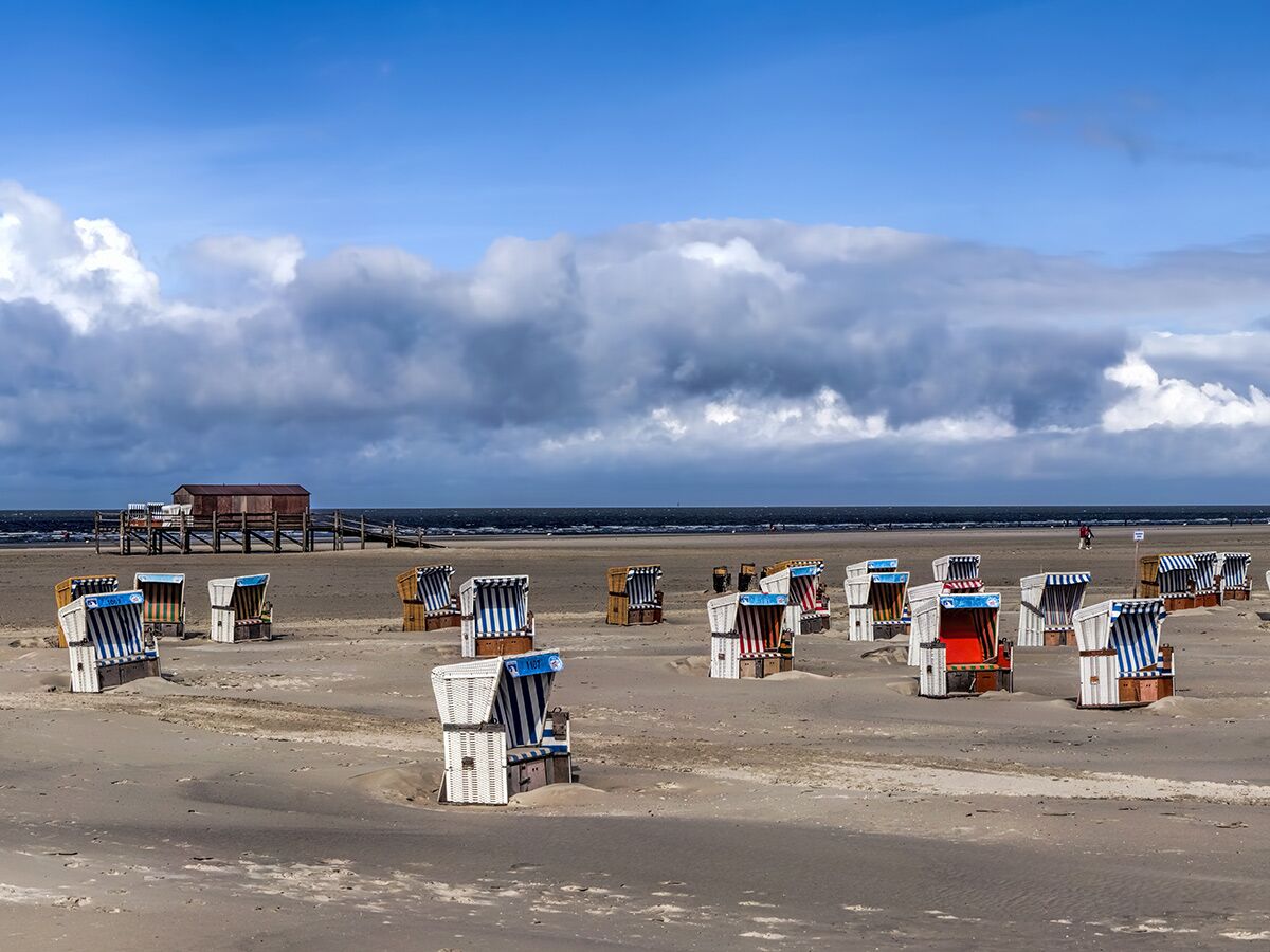 Zahlreiche leere Strandkörbe stehen auf weitem Sandstrand unter bewölktem Himmel. Meer und Pfahlbau im Hintergrund sichtbar.