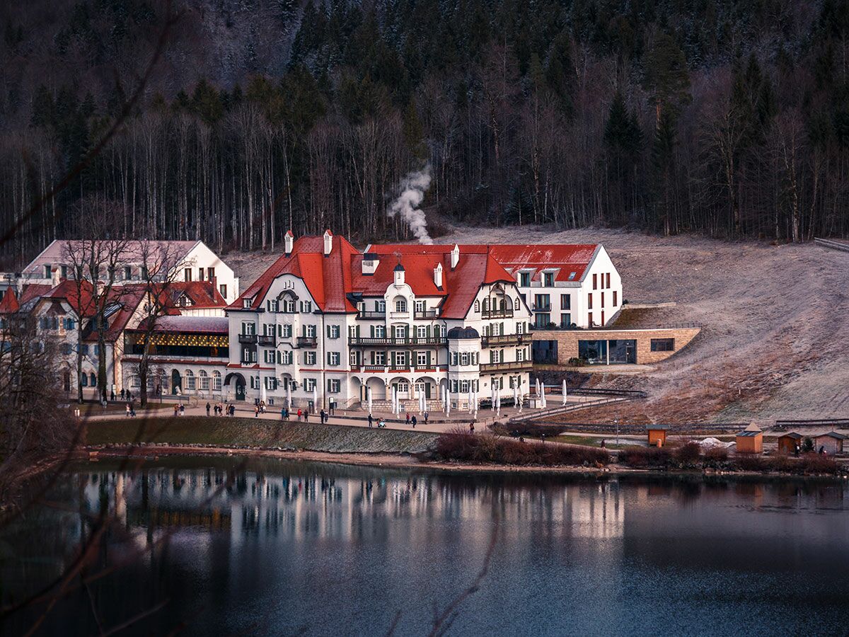 Großes Hotel mit roten Dächern und weißen Fassaden am Seeufer, umgeben von Wald und frostigen Hügeln, mit Menschen am Weg.