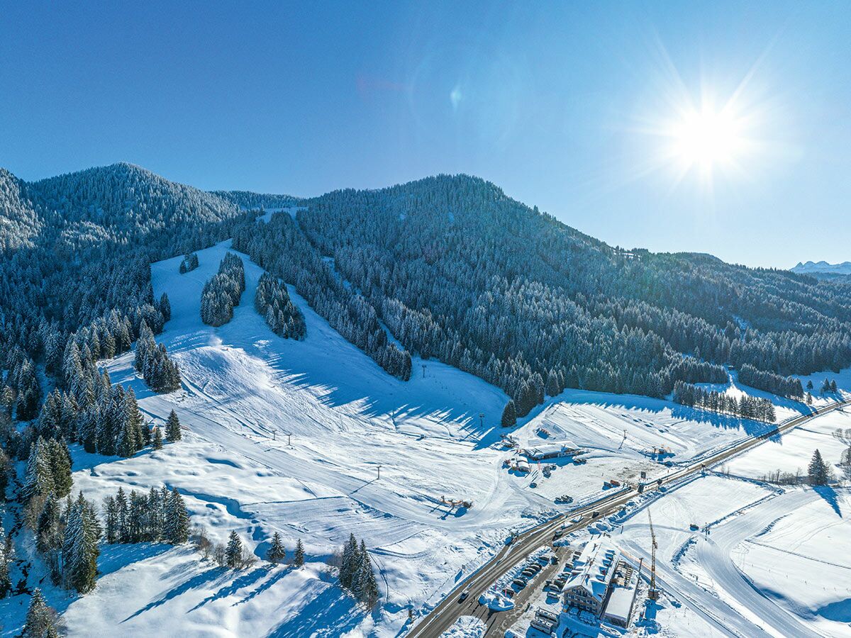 Luftaufnahme eines verschneiten Skigebiets in den Alpen mit Skipisten, Nadelbäumen, einer Straße und Gebäuden unter strahlender Wintersonne.