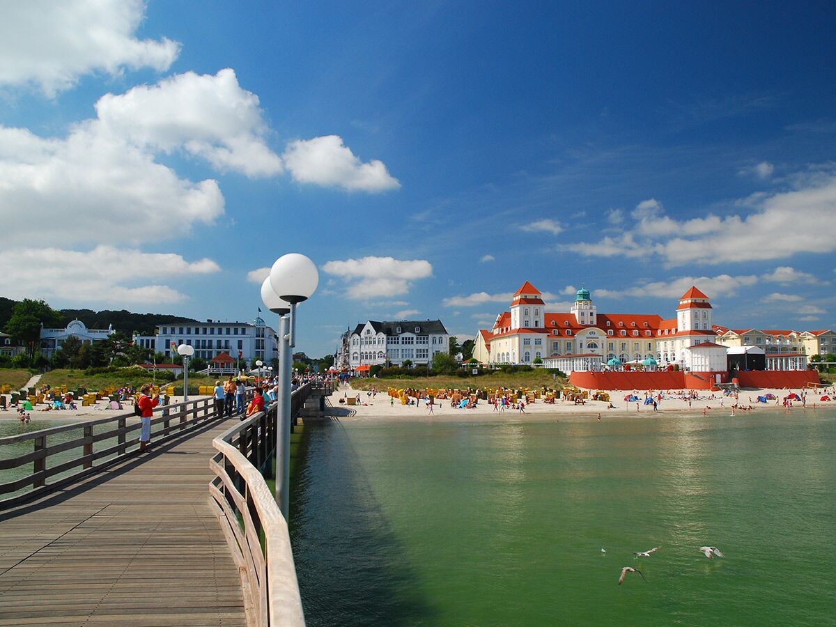 Lange Holzseebrücke mit Spaziergängern am Strand von Binz auf Rügen. Im Hintergrund prächtige Hotels an der Ostseeküste unter blauem Himmel.
