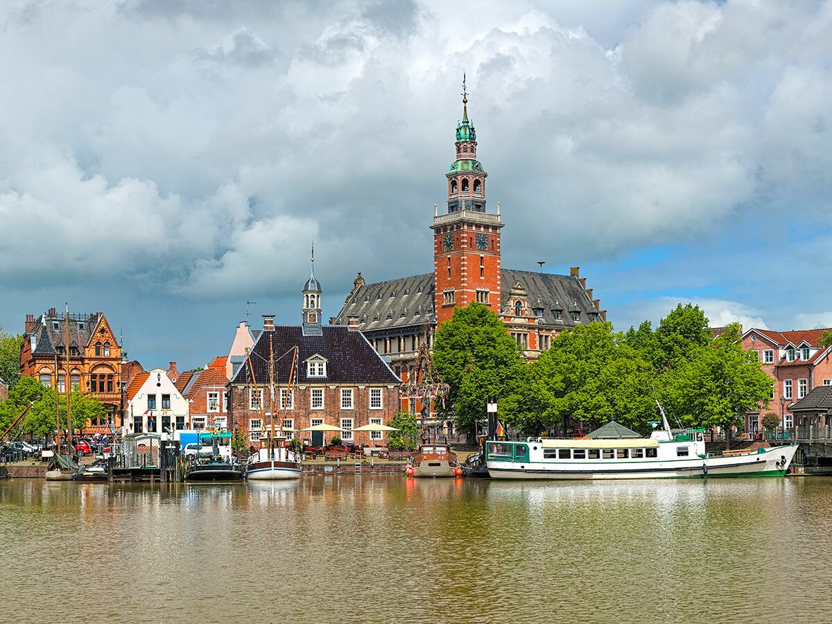 Historische Hafenstadt Leer, Deutschland, mit imposantem Rathaus, bunten Gebäuden, Booten am Kanal und grünen Bäumen unter bewölktem Himmel.