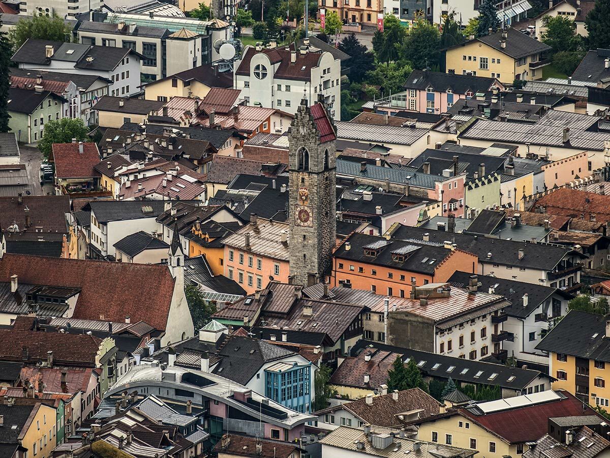Luftbild einer historischen Alpenstadt mit vielen bunten Häusern, roten Dächern und einem zentralen Kirchturm mit Uhr.