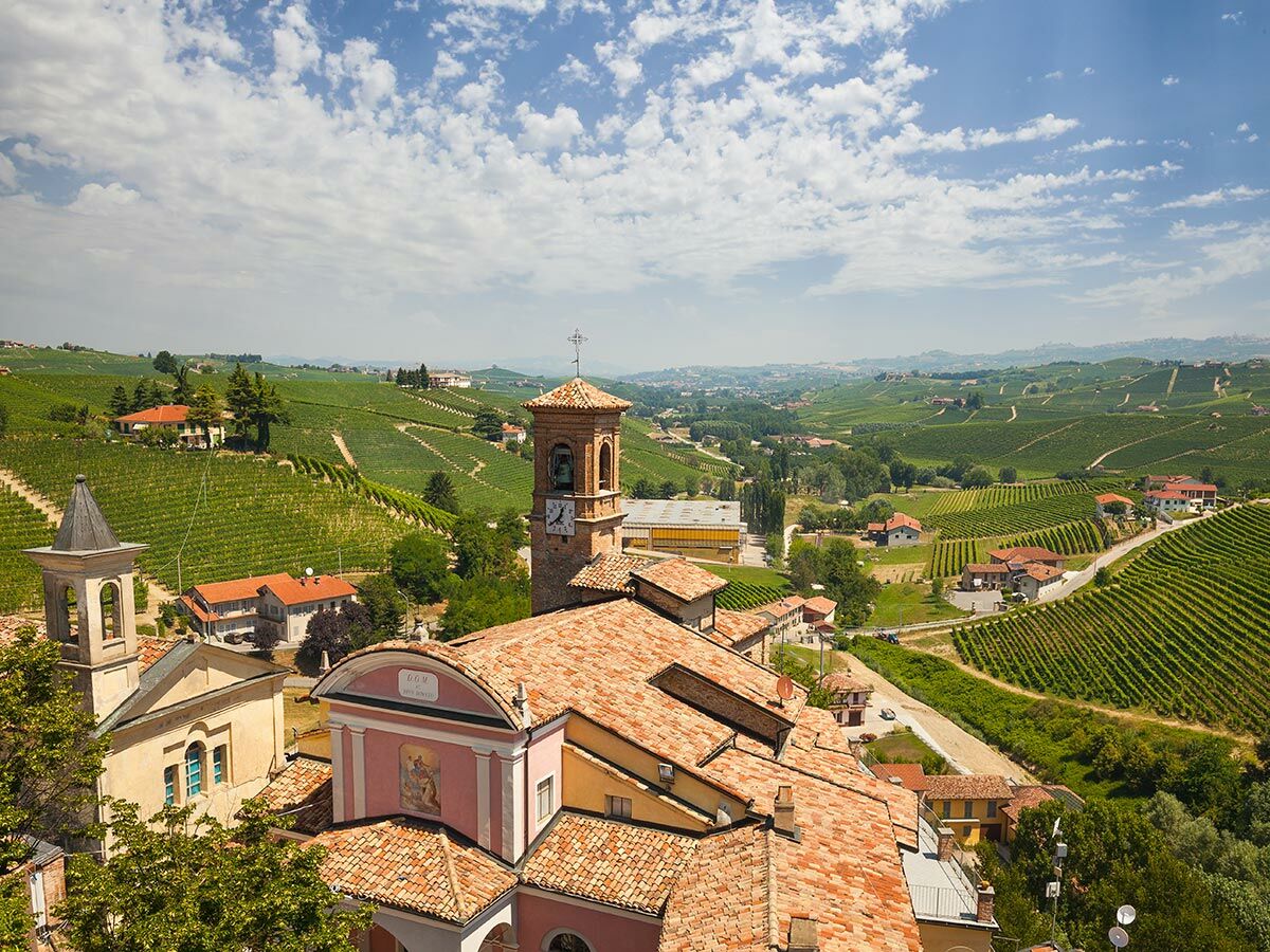 Panoramablick auf ein italienisches Dorf im Piemont, umgeben von grünen Weinbergen, mit Kirchturm, roten Dächern und Hügellandschaft unter blauem Himmel.