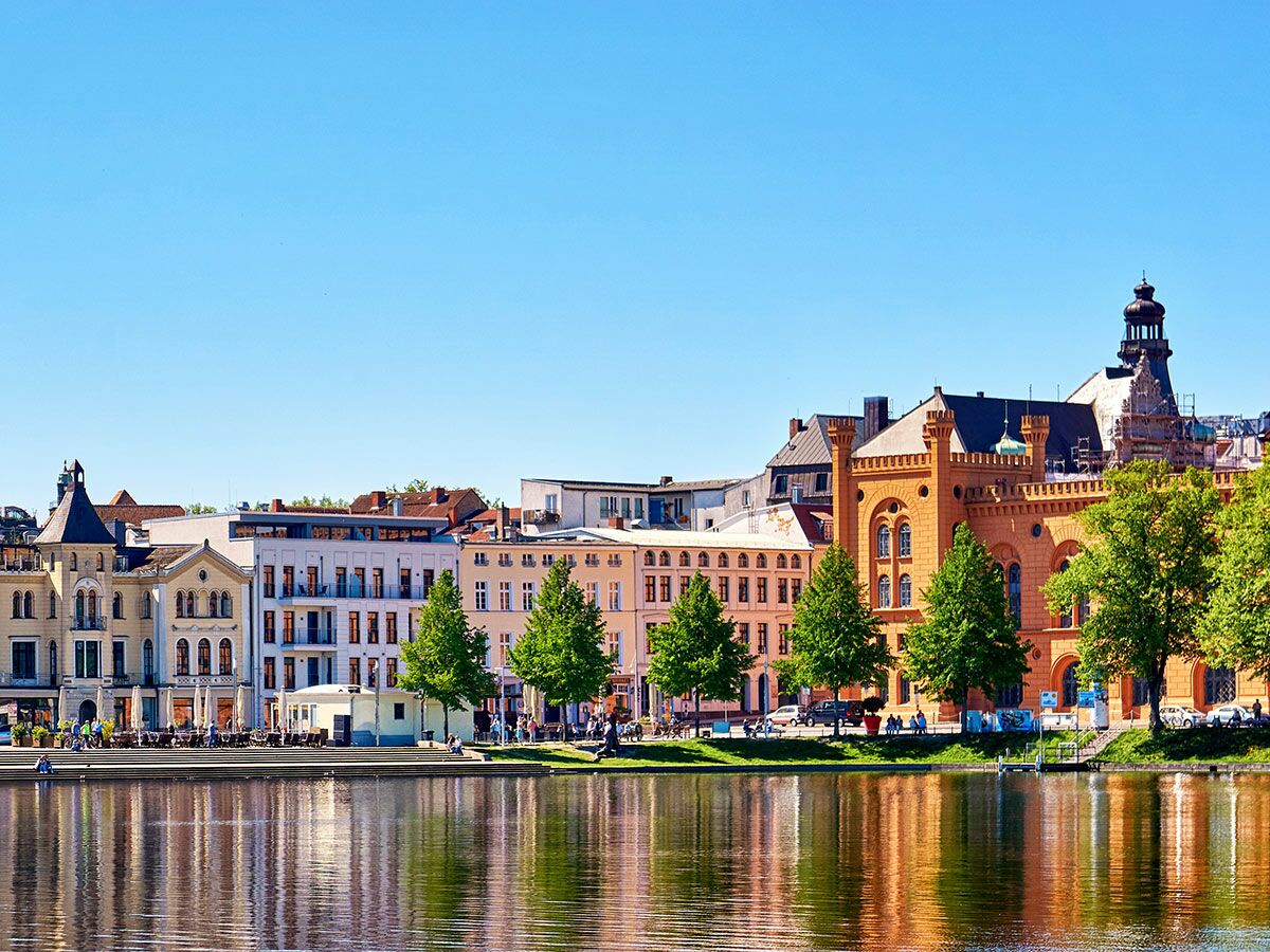 Stadtansicht von Schwerin am Pfaffenteich mit historischen Gebäuden, Menschen und Reflexionen im Wasser an einem sonnigen Tag.