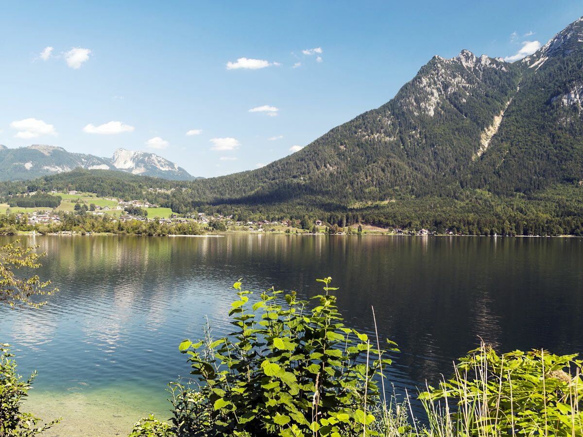 Idyllischer Bergsee in den Alpen mit Dorf am Ufer, umgeben von Wäldern und majestätischen Bergen unter blauem Himmel.