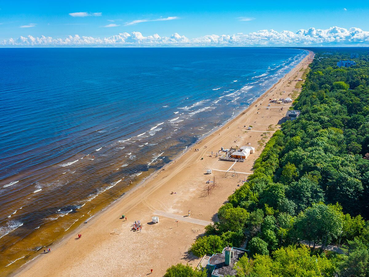 Drohnenansicht langer Sandstrand mit Menschen, Meer, blauem Himmel und Wald an sonnigem Sommertag.