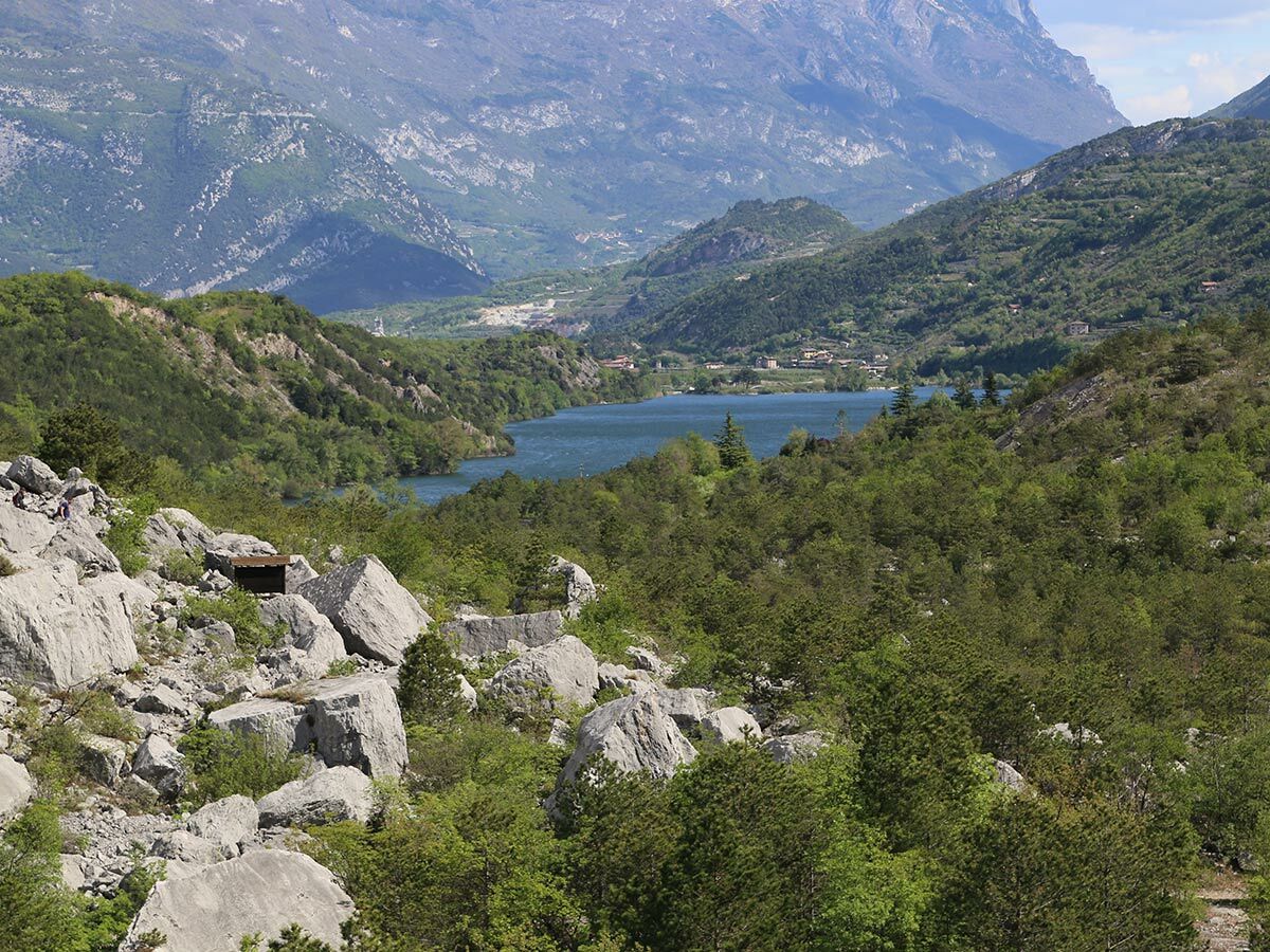 Panoramablick auf einen blauen Bergsee, umgeben von grünen Hügeln, bewaldeten Bergen und einem alten Bunker im Fels.
