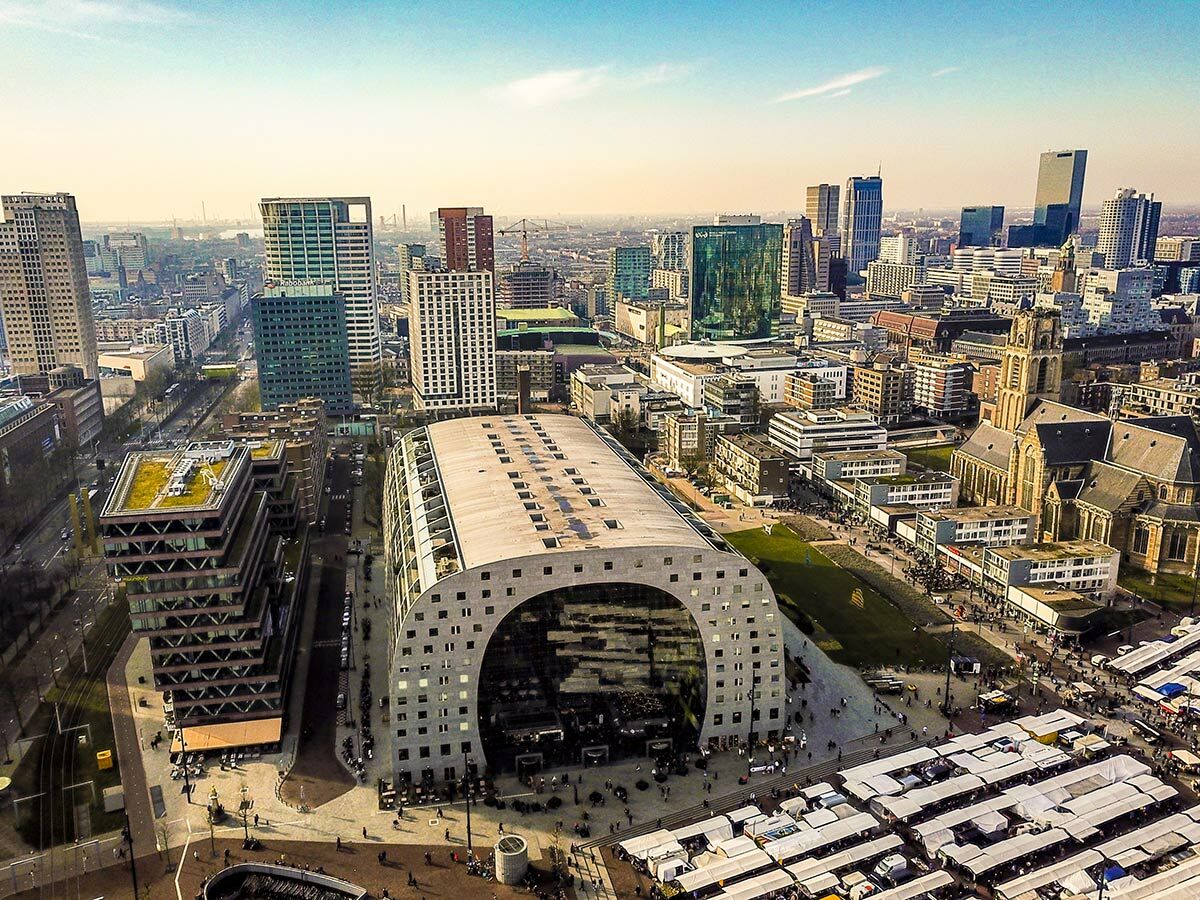 Panoramablick auf die moderne Stadt Rotterdam mit der ikonischen Markthal, hohen Gebäuden, der St. Laurenskerk und einem lebhaften Markt.