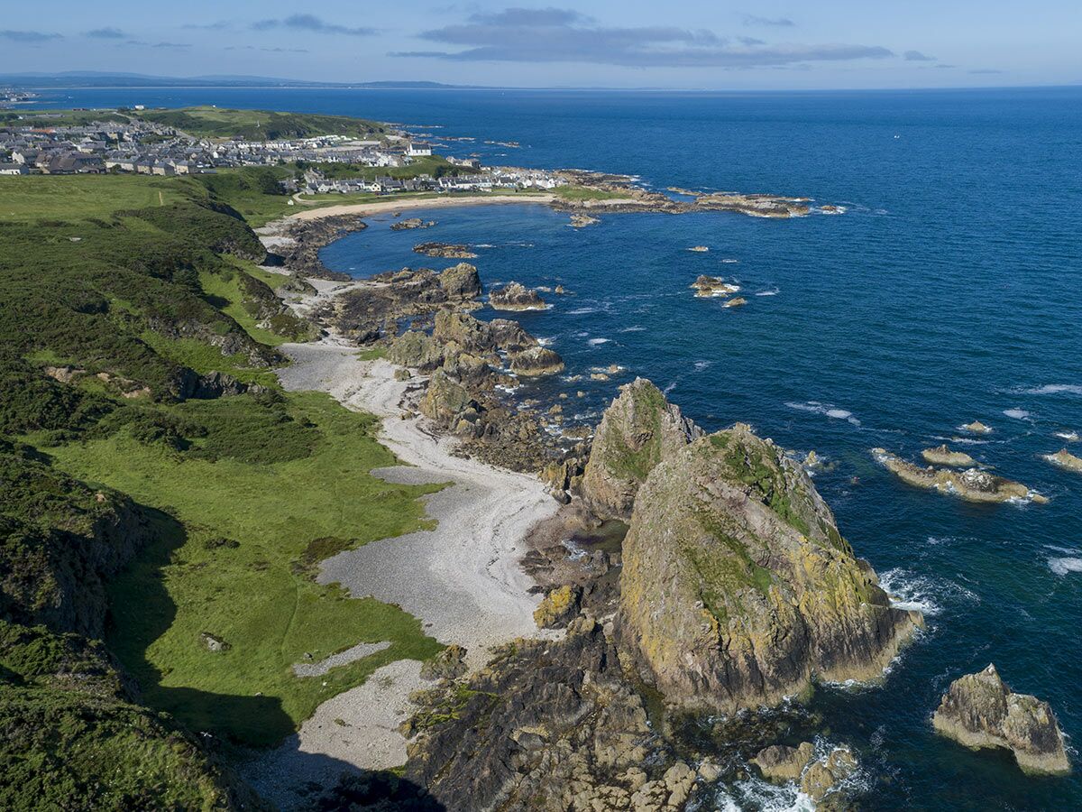 Luftaufnahme einer schottischen Küstenlandschaft mit einem Küstendorf, grünen Klippen, Felsformationen und ruhigem Meer.
