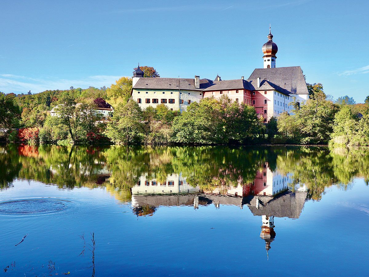 Malerische Ansicht eines farbenfrohen historischen Klosters mit Zwiebelturm-Kirche, das sich in einem ruhigen See spiegelt, umgeben von Herbstbäumen.