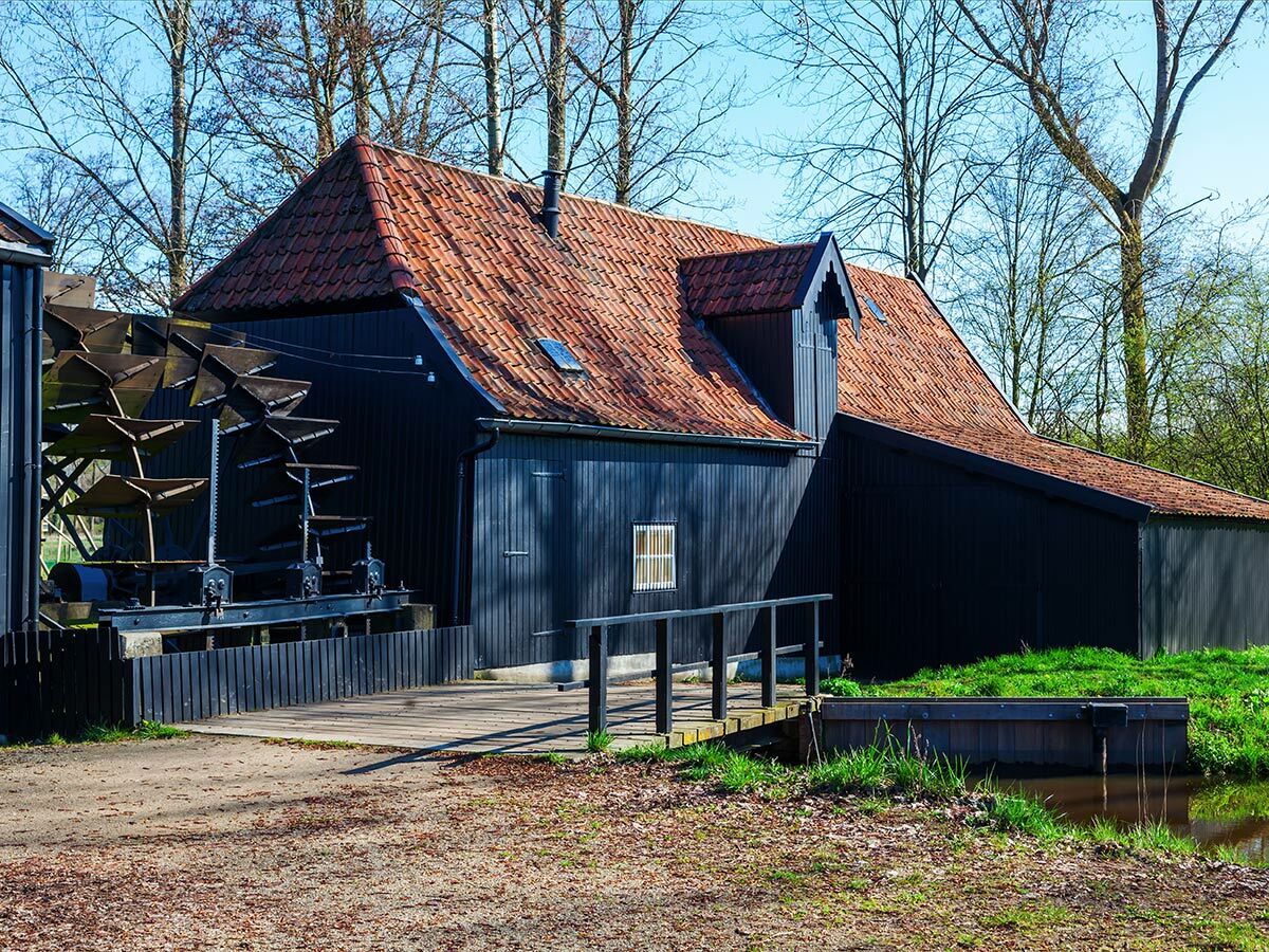 Eine historische Wassermühle aus dunklem Holz mit großem Wasserrad und rotem Ziegeldach steht am Ufer eines kleinen Baches.
