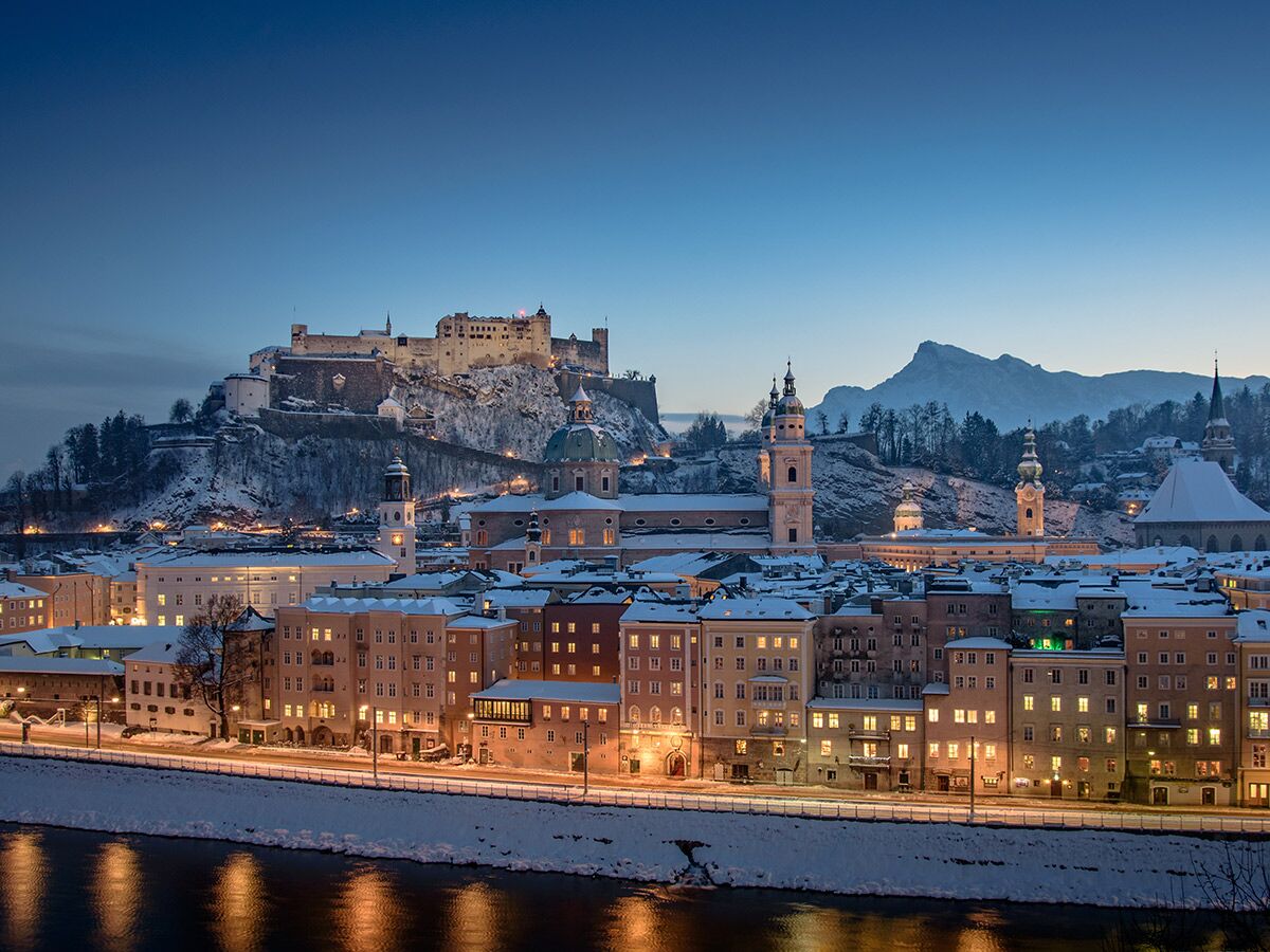 Historisches Salzburg im Winter bei Dämmerung, verschneite Dächer, beleuchtete Gebäude, Festung Hohensalzburg über der Salzach.