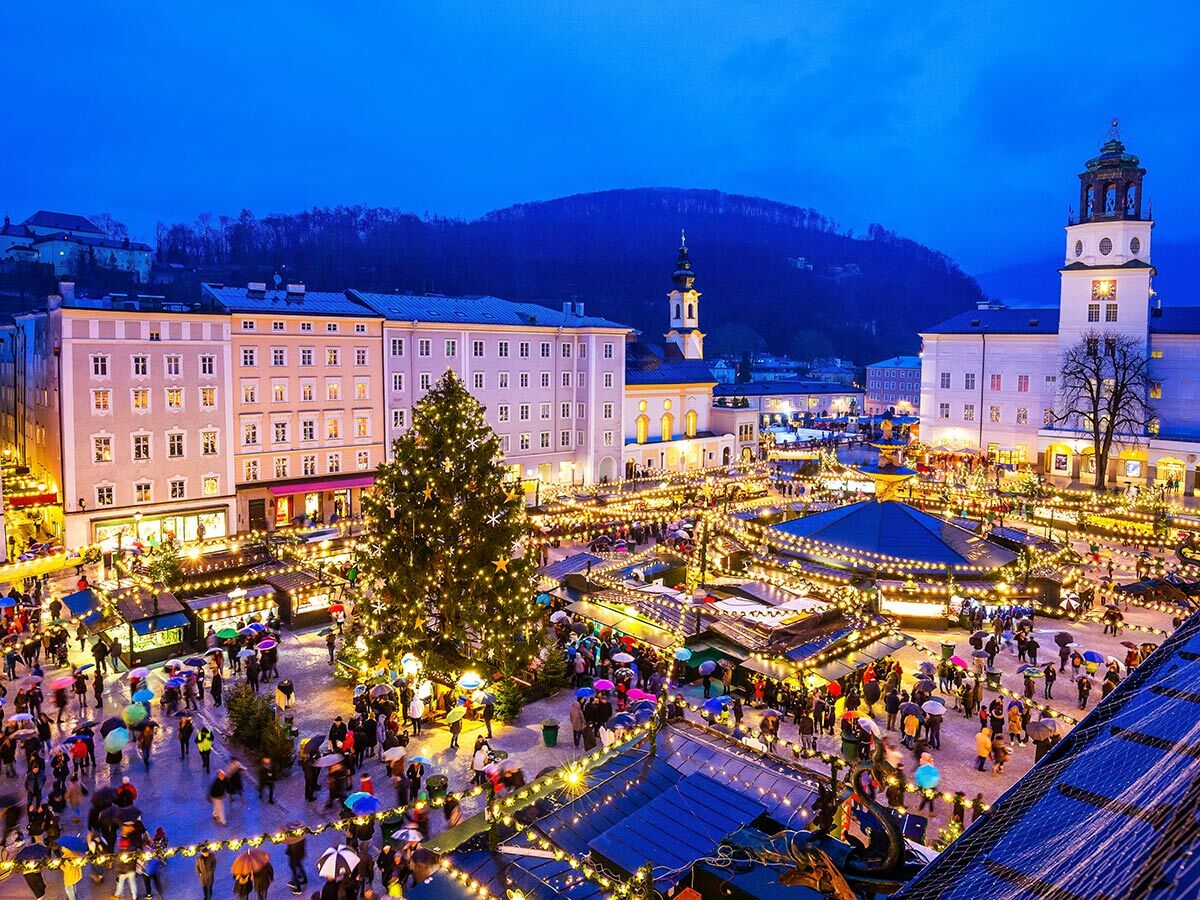 Beleuchteter Salzburger Christkindlmarkt am Abend mit großem Weihnachtsbaum, Marktständen und vielen Menschen unter Regenschirmen.