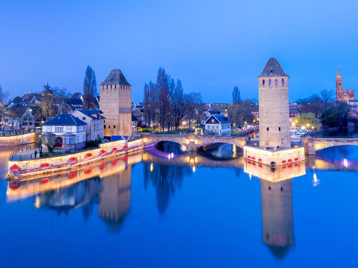 Historische Ponts Couverts in Strasbourg bei Blaue Stunde. Zwei beleuchtete Türme und Brücken spiegeln sich im Fluss Ill.