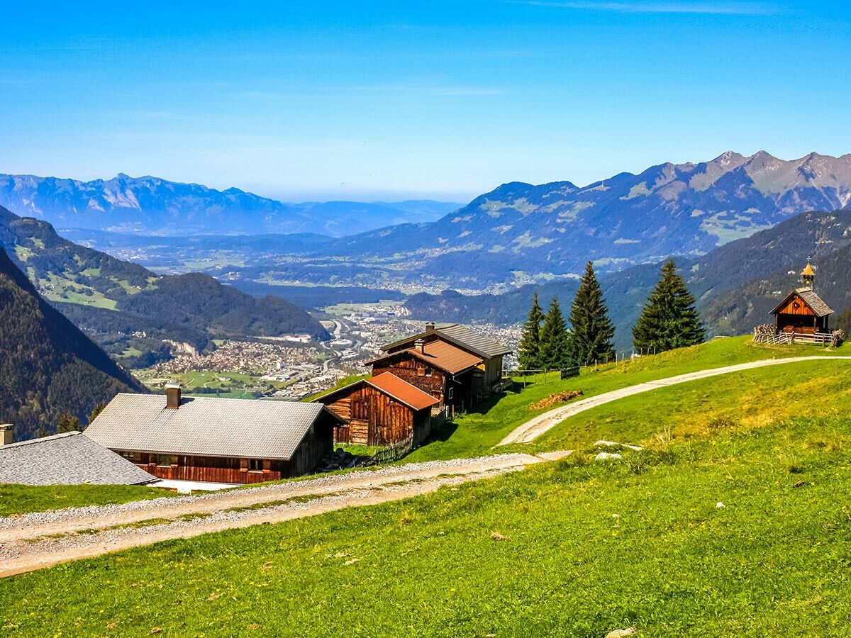 Panoramablick auf ein Alpentall mit traditionellen Holzhäusern, einer kleinen Kapelle, grünen Wiesen und fernen Bergen unter blauem Himmel.