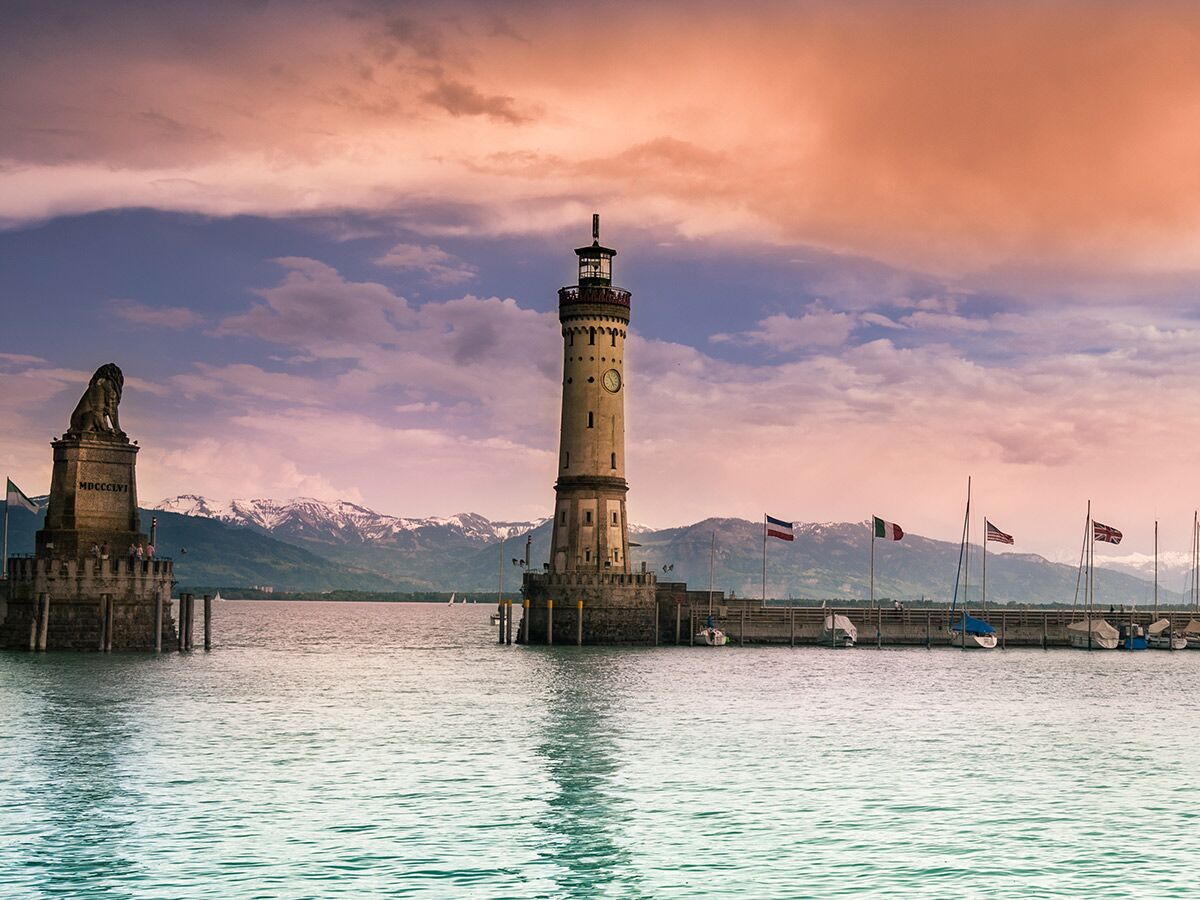 Panoramablick auf den Lindauer Hafen mit Leuchtturm, Löwenstatue und verschneiten Alpen bei Sonnenuntergang.