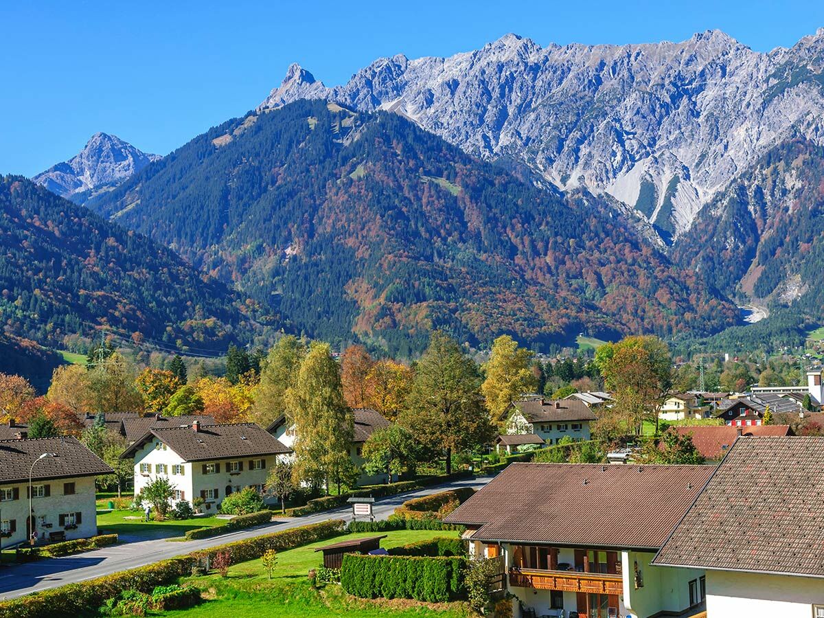 Malerisches Dorf in den Alpen im Herbst mit bunten Bäumen und majestätischen Bergen unter blauem Himmel.