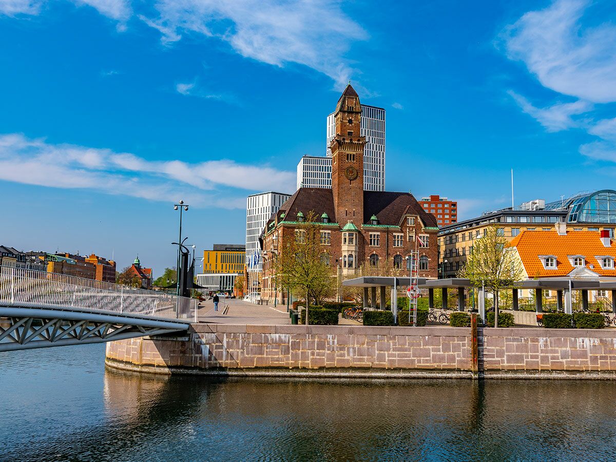 Panorama von Malmö mit der historischen Stadtbibliothek, modernen Gebäuden, einem Kanal, einer Brücke und Passanten unter blauem Himmel.