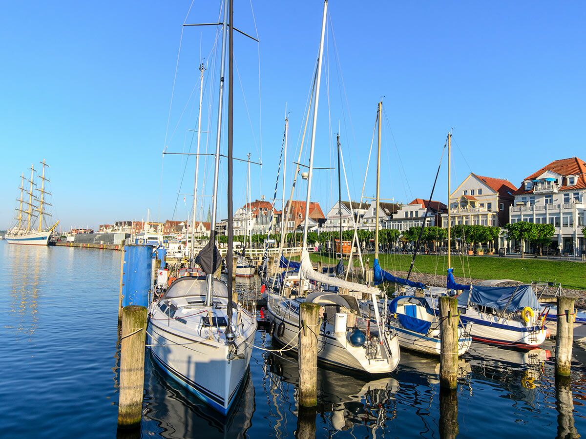 Blick auf einen sonnigen Hafen mit modernen Segelbooten, Anlegestellen und einem majestätischen Tallship, umgeben von Stadthäusern.