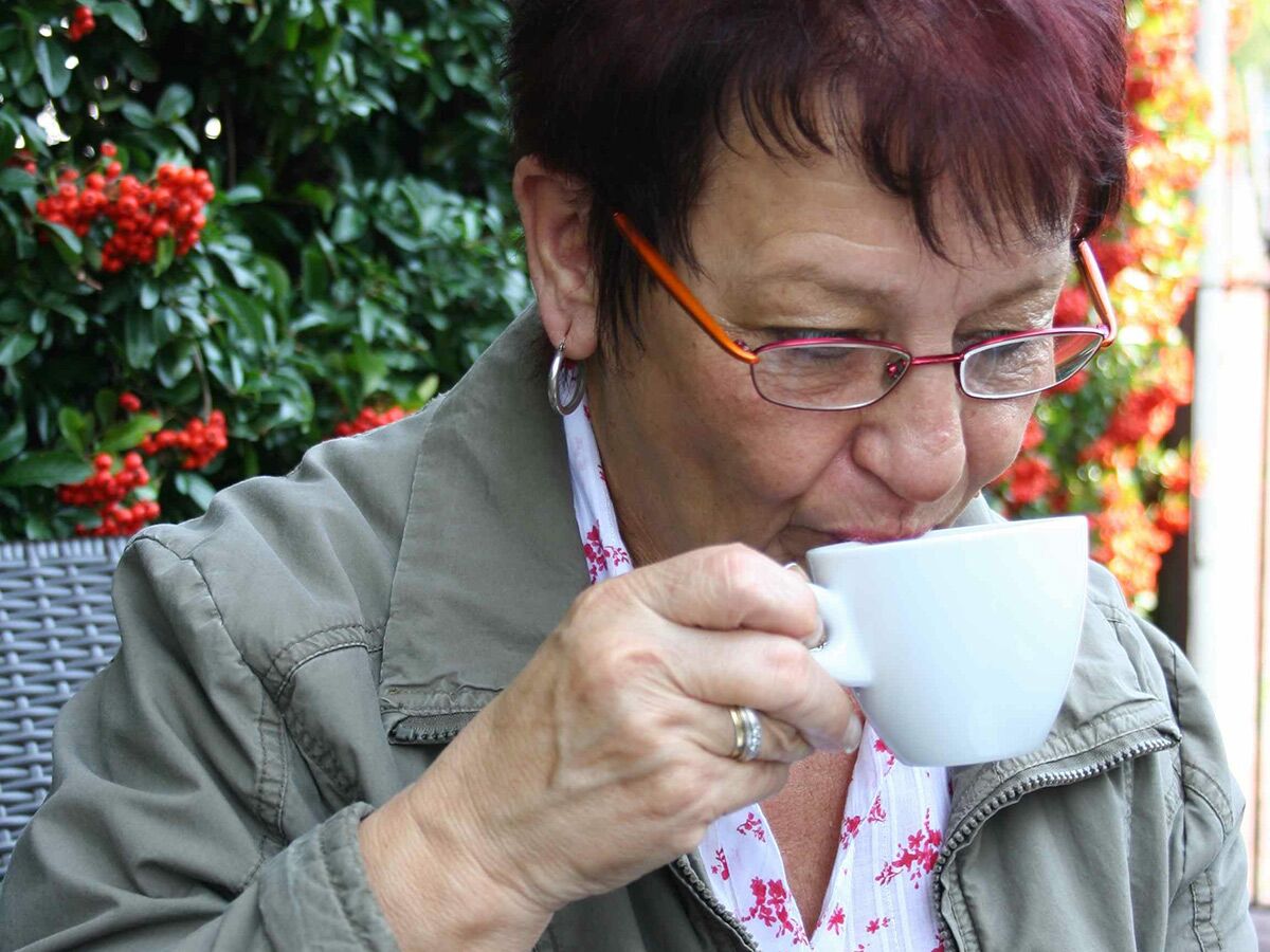 Nahaufnahme einer Frau mit Brille, die draußen aus einer weißen Tasse trinkt, umgeben von grünen Blättern und roten Beeren.