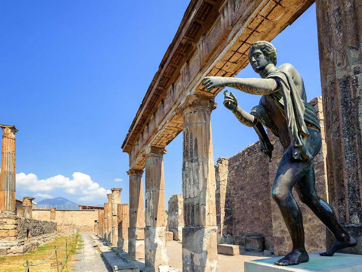 Bronze-Statue des Apollo mit Blick auf antike Säulen und Ruinen von Pompeji unter blauem Himmel mit dem Vesuv im Hintergrund.