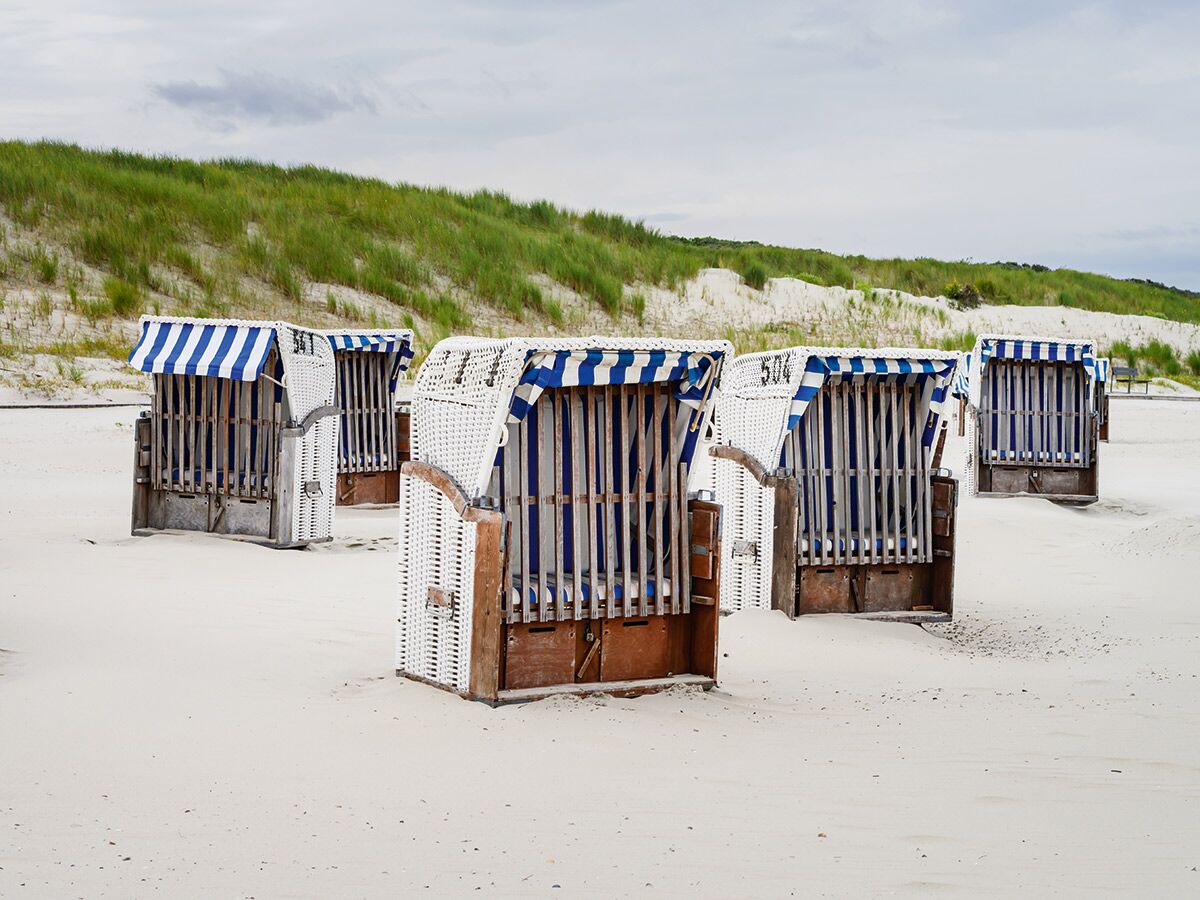 Mehrere blau-weiß gestreifte Strandkörbe stehen auf einem sandigen Strand vor grünen Dünen unter bewölktem Himmel am Meer.
