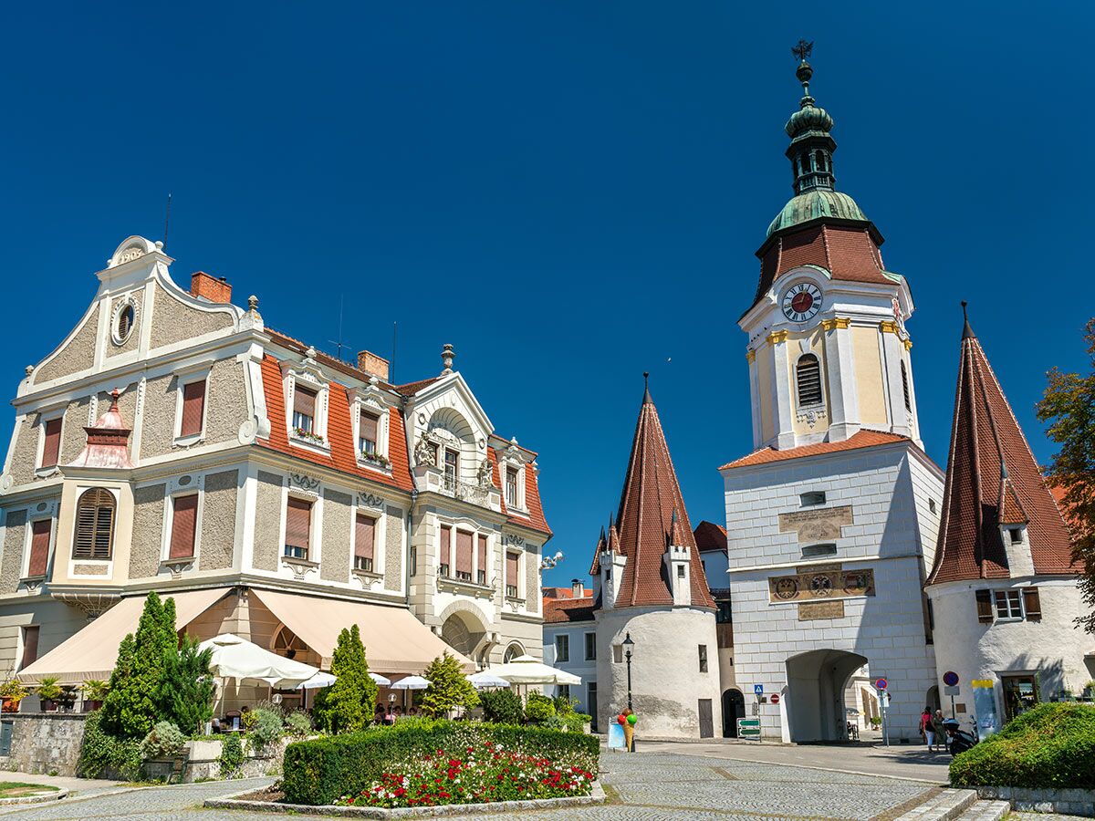 Historischer Stadtplatz von Wels, Österreich, mit barockem Uhrturm, alten Häusern und einem belebten Straßencafé unter blauem Himmel.