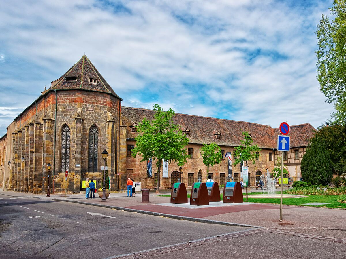 Historisches Unterlinden Museum in Colmar, Elsass, mit Dominikanerkirche, Straße und Platz unter bewölktem Himmel.