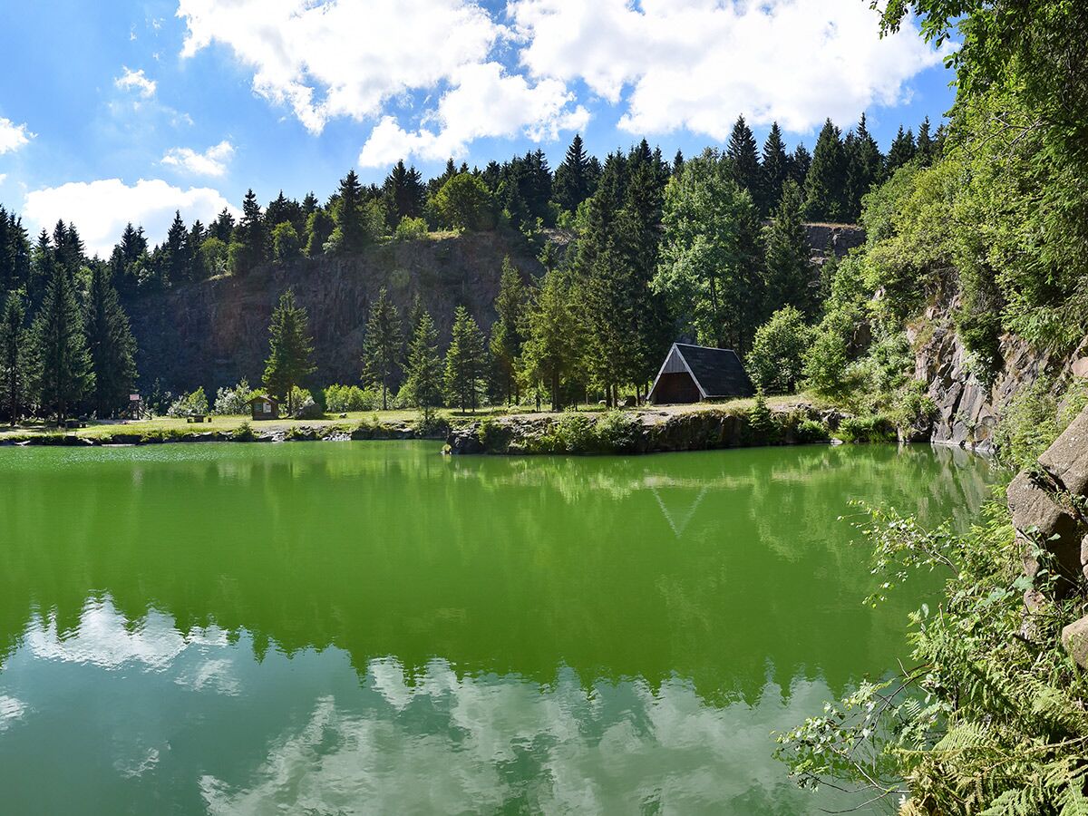 Grüner Steinbruchsee, umgeben von Wäldern, Felsklippen und einer Holzhütte unter blauem Himmel.