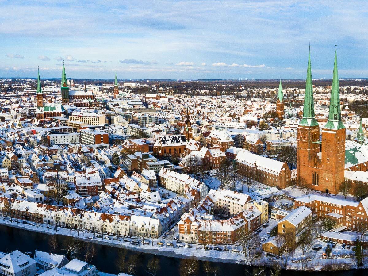 Luftaufnahme von Lübeck im Winter mit schneebedeckten Dächern und dem Dom mit seinen grünen Spitzen unter blauem Himmel.