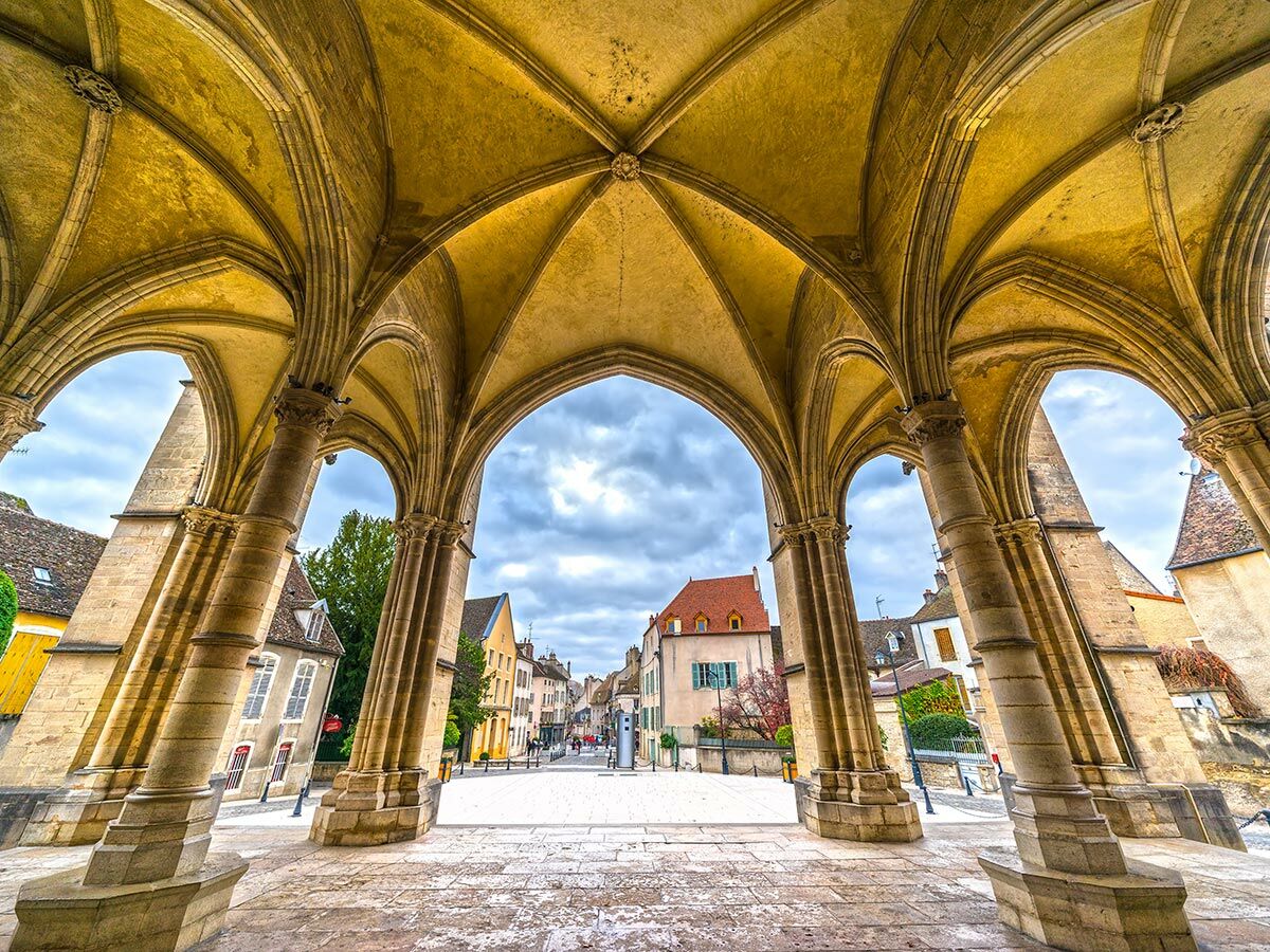 Blick durch kunstvolle gotische Steinbögen auf eine historische europäische Stadtstraße mit alten Gebäuden unter bewölktem Himmel.