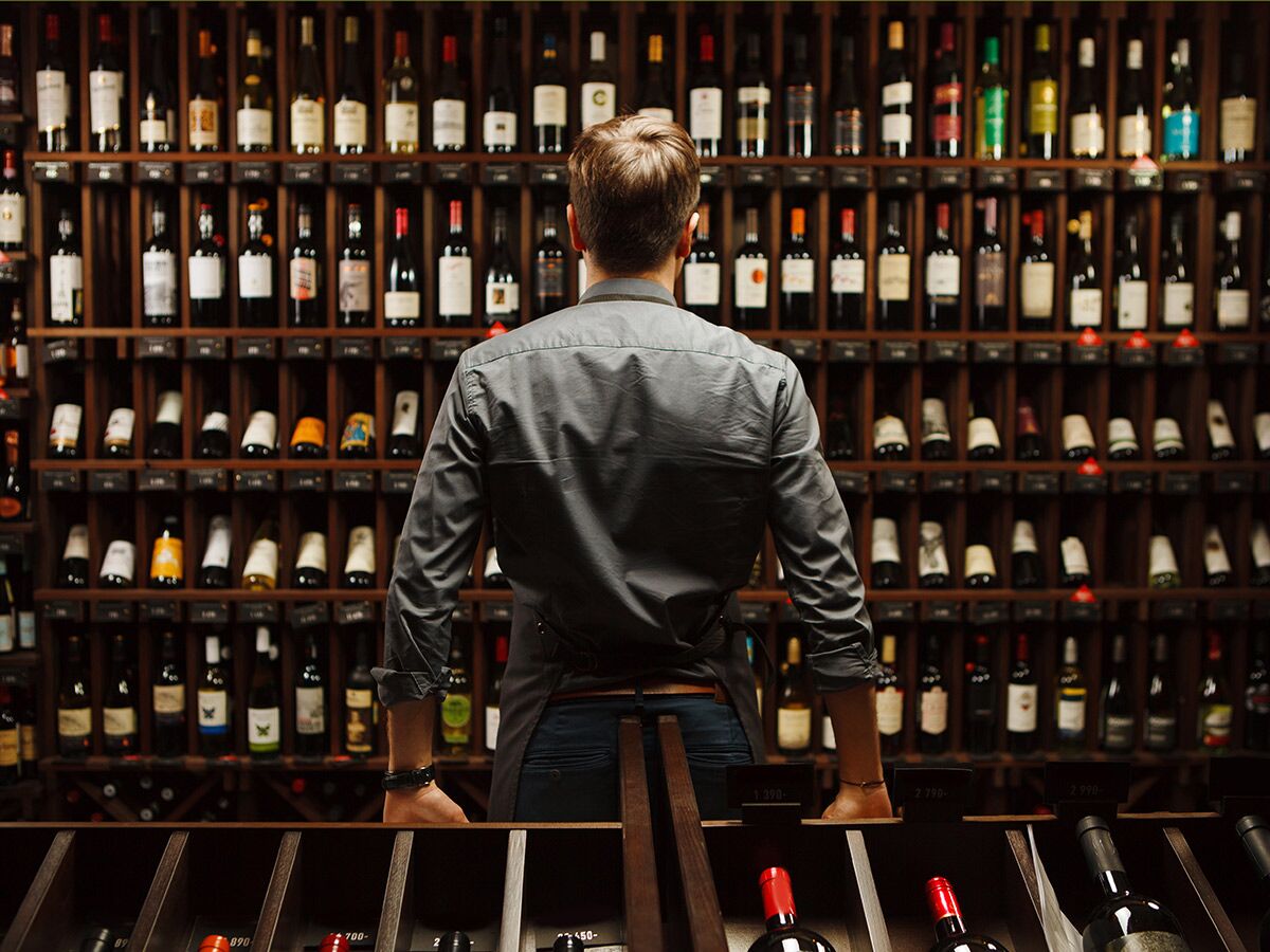 Mann von hinten in einem Weinladen, blickt auf eine große Auswahl an Weinflaschen in Holzregalen mit Preisschildern.
